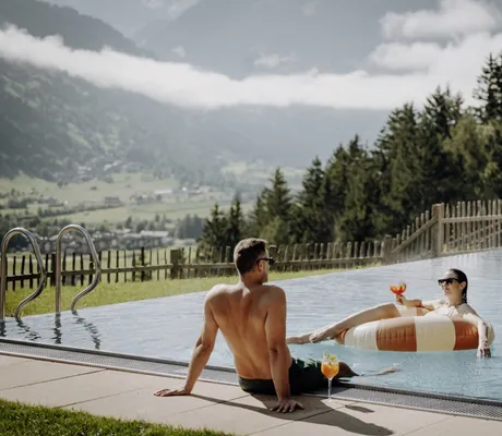 Mann am Outdoorpool mit Cocktail schaut Frau in gestreiftem Schwimmreifen im Pool im Das.Goldberg zu