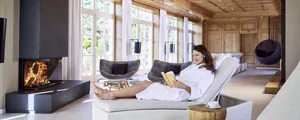 Frau in weißem Bademantel liest auf der Liege vor Kamin im Interalpen-Hotel Tyrol und liest ein Buch