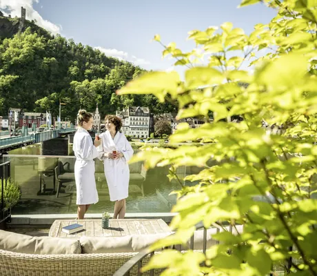 zwei Frauen in weißen Bademänteln unterhalten sich auf der Spa-Terrasse des Moselschlösschen Spa & Resort mit Blick auf die Moselbrücke