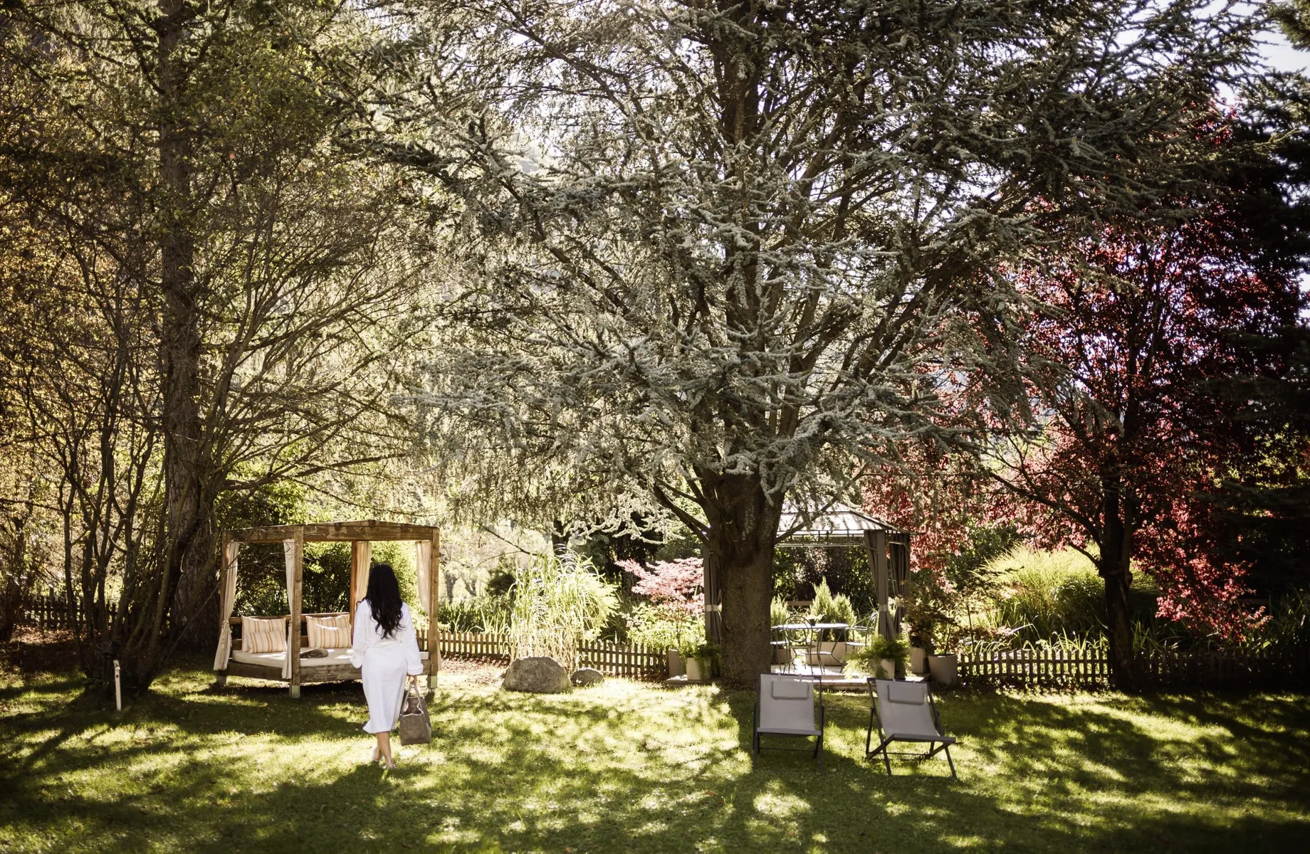 Frau in weißem Bademantek und Badtasche geht im Park des Romantik Hotel Stafler zu einem Daybed