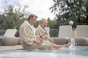 Best Ager-Paar sitzt mit beigen Bademänteln am Beckenrand des Pools im Romantischer Winkel und trinkt Rosé
