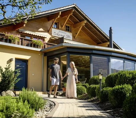 Frau in bodenlangem Kleid und Mann mit Brille gehen Hand in Hand den Weg im Wellnessgarten des Salzano entlang