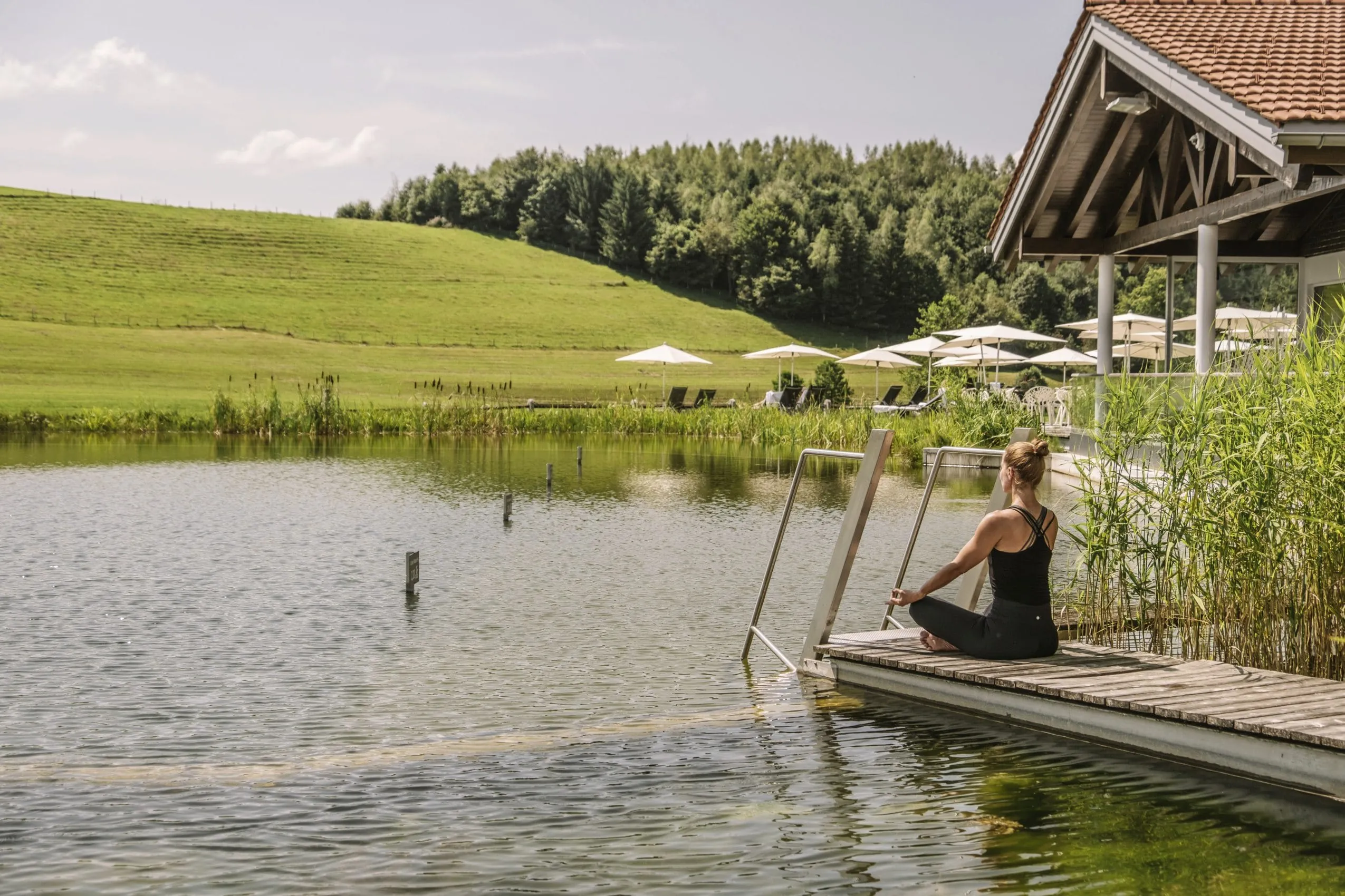 Frau in schwarzem Gym-Outfit macht Yoga auf dem Steg am Natursee in Haubers Naturresort