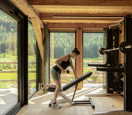 Frau in Sportkleidung beugt sich über eine Hantelbank im Fitnessraum der Südtirol Chalets Valsegg, im Hintergrund große Fenster mit Blick auf eine bewaldete Landschaft