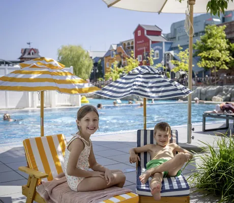 Zwei Kinder auf kleinen Kinderliegen am Pool. Im Hintergrund ist ein Pool und bunte Gebäude des Krønasår – The Museum-Hotel zu sehen.