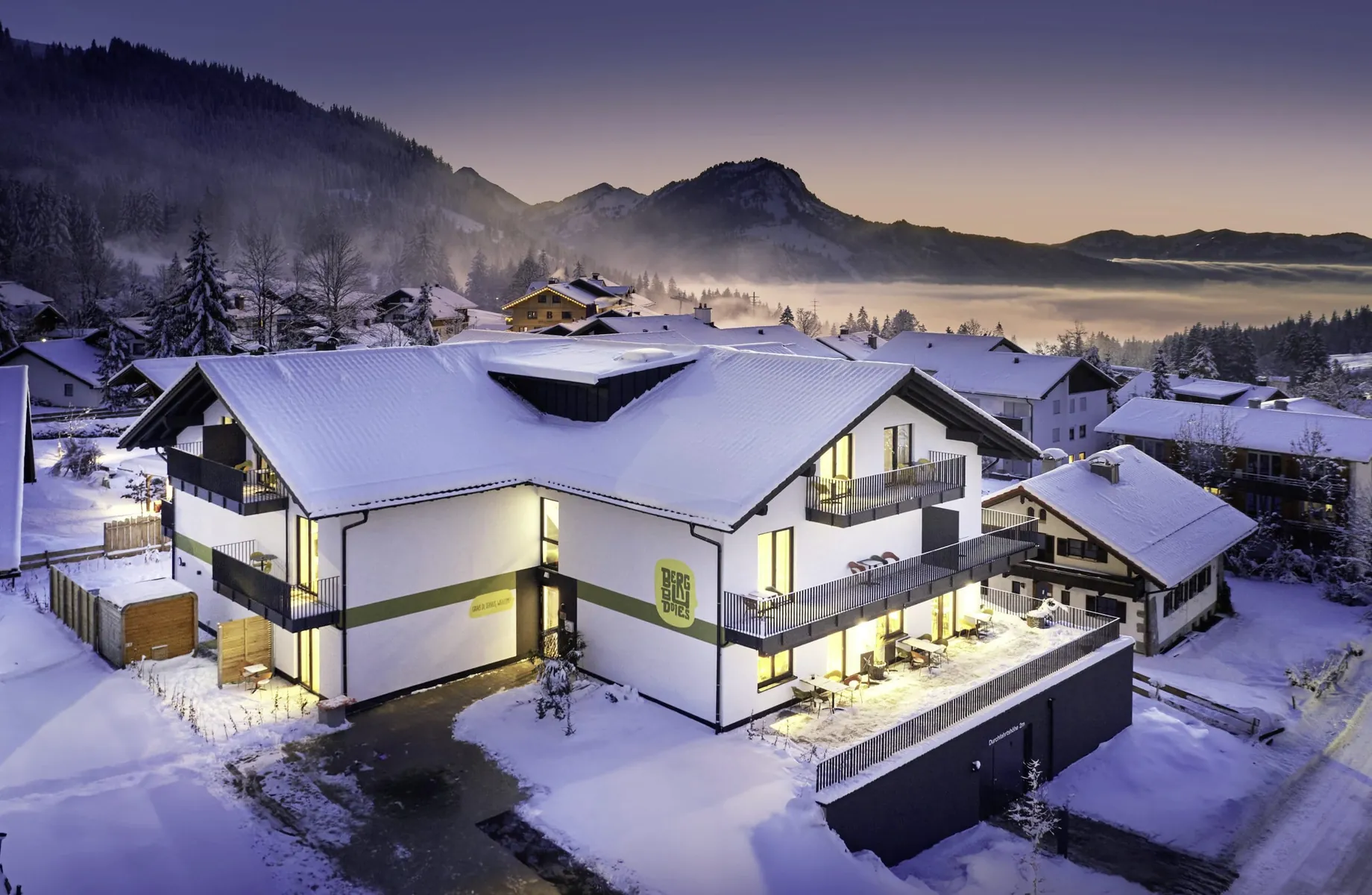 Blick aus der Vogelperspektive auf das eingeschneite und beleuchtete BergBuddies-Hotel im Winter