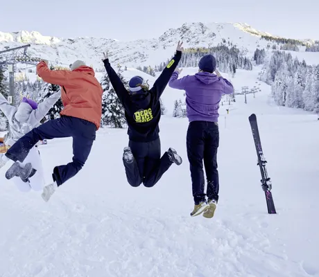 Vier junge Menschen machen vor Freude einen Luftsprung im Schnee, im Hintergrund das Skigebiet von Oberjoch