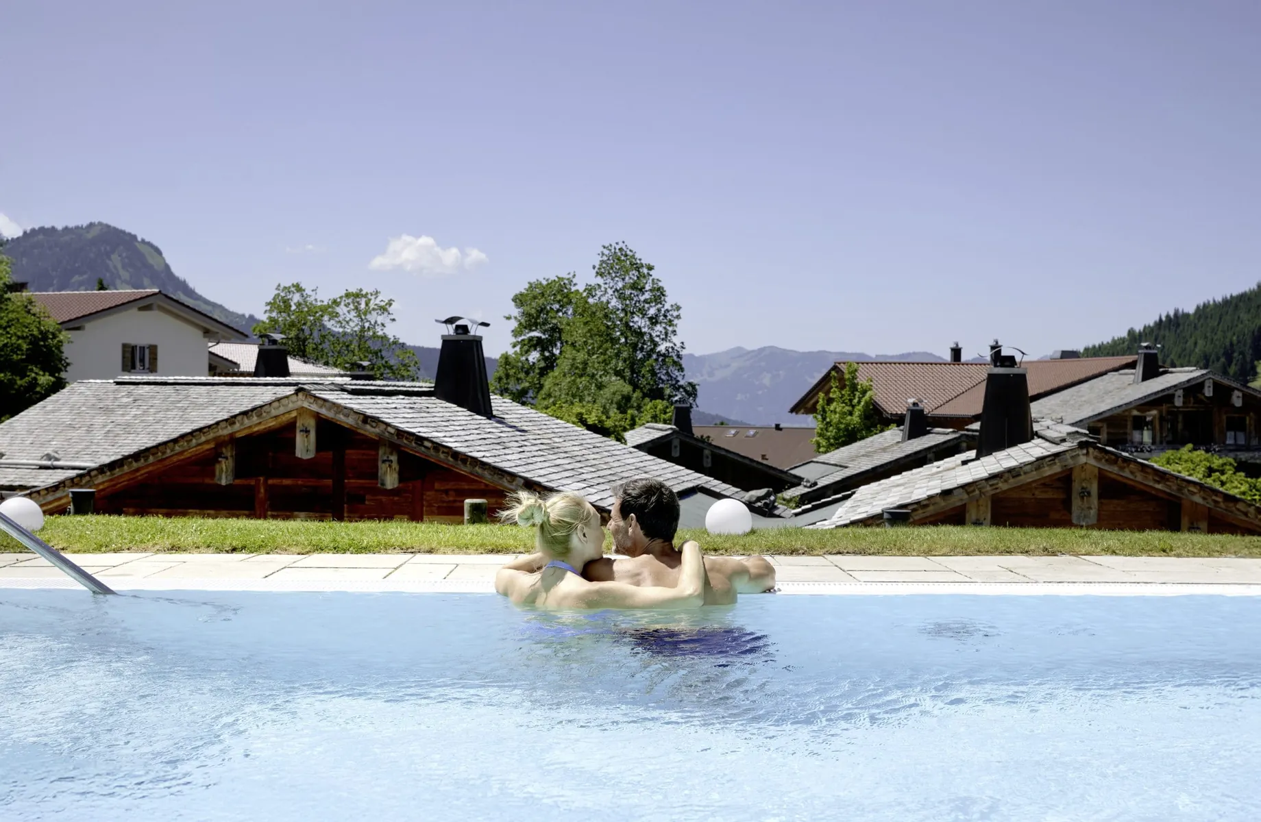 Paar steht umarmt im Outdoorpool des Panoramahotel Oberjoch und schaut auf das Chaletdorf