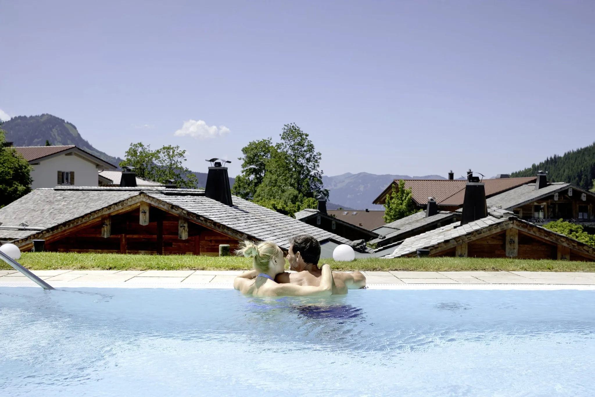 Paar steht umarmt im Outdoorpool des Panoramahotel Oberjoch und schaut auf das Chaletdorf
