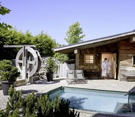 Paar in Bademänteln kommt aus der Blockhaussauna am Tauchbecken mit Mühlrad im Panoramahotel Oberjoch