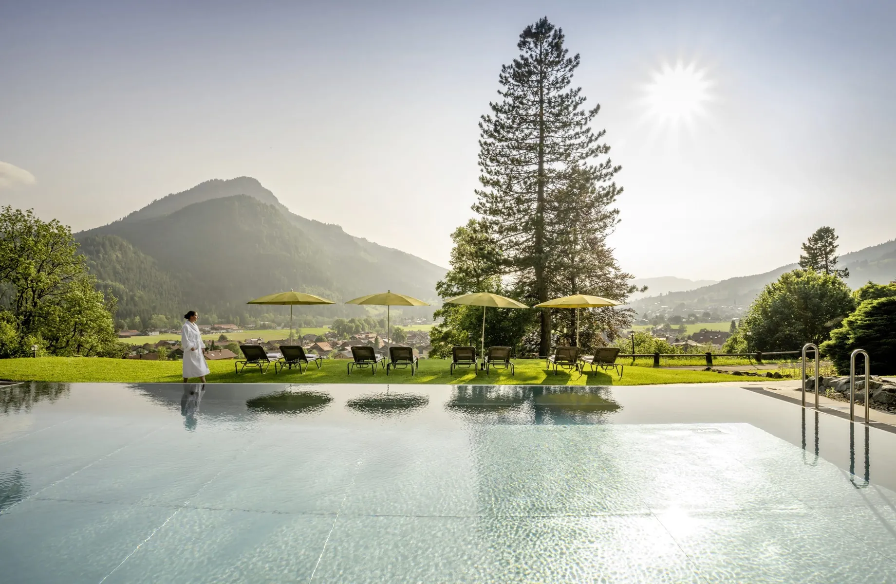 Frau in weißem Bademantel läuft am Outdoorpool des Hotel Prinz-Luitpold-Bad mit gelben Sonnenschirmen entlang