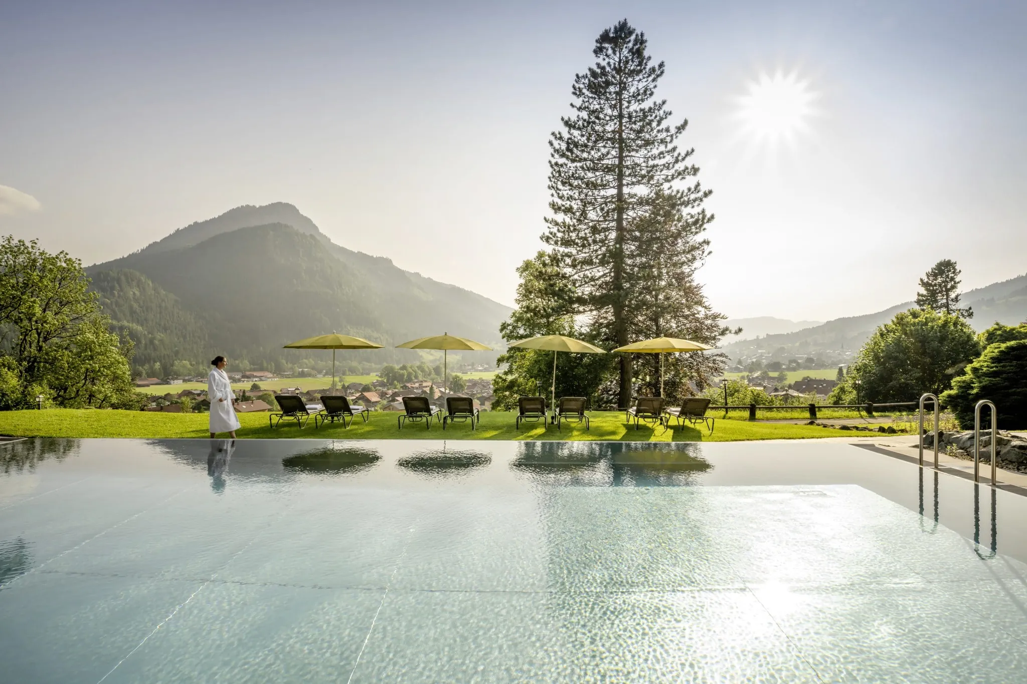 Frau in weißem Bademantel läuft am Outdoorpool des Hotel Prinz-Luitpold-Bad mit gelben Sonnenschirmen entlang