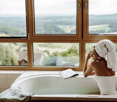 Frau in weißem Badanzug und mit weißem Handtuchturban sitzt an der Wanne am Fenster im Thula Wellnesshotel
