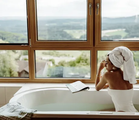Frau in weißem Badanzug und mit weißem Handtuchturban sitzt an der Wanne am Fenster im Thula Wellnesshotel
