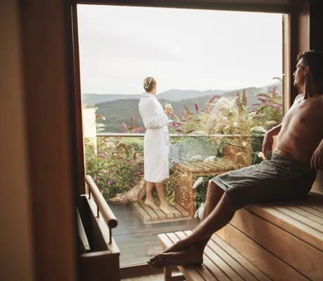 Mann in dunklem Frottetuch sitzt in der Sauna und schaut auf Frau draußen in weißem Bademantel in Thula Wellnesshotel