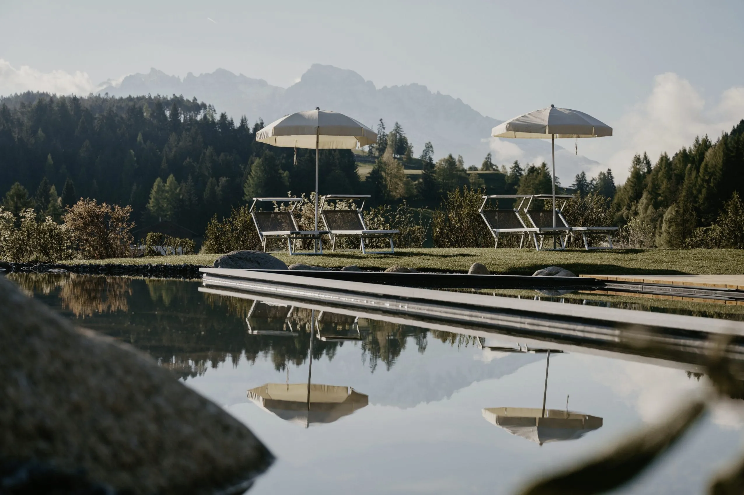 Outdoorpool in Naturteich integriert mit Liegest&uuml;hlen und Sonnenschirmen vor Bergpanorama im Naturhotel Pf&ouml;sl