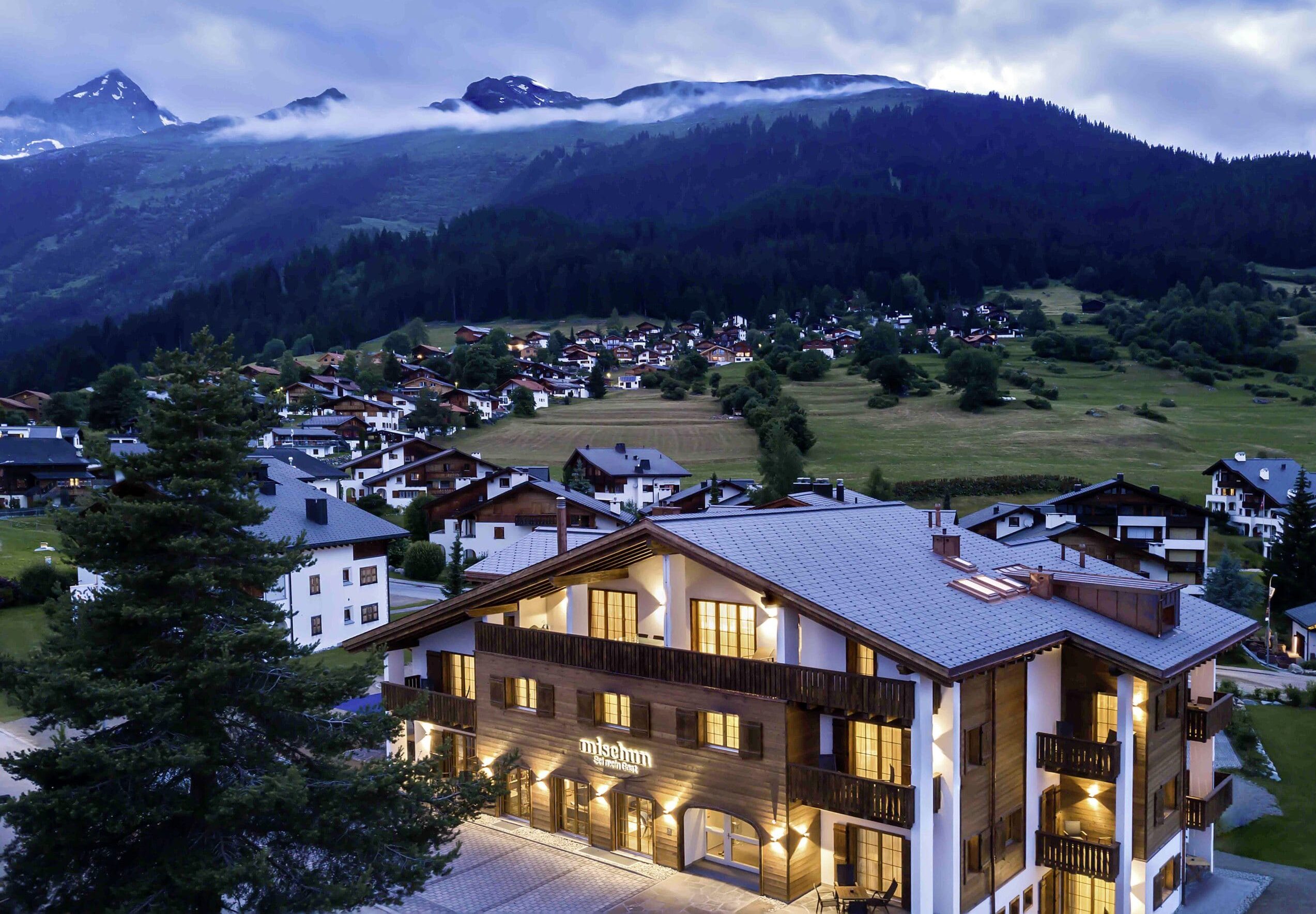 Beleuchtete Holzfassade des Hotel Mischun in einem Bergdorf, im Hintergrund bewaldete Berge unter bewölktem Himmel