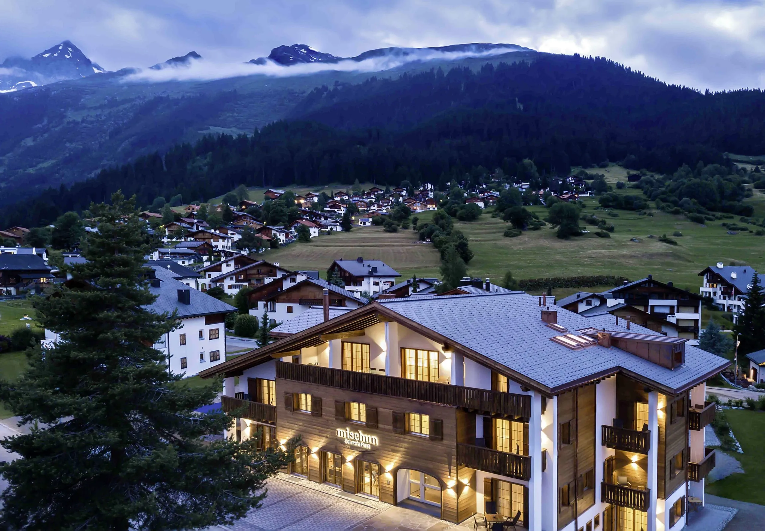 Beleuchtete Holzfassade des Hotel Mischun in einem Bergdorf, im Hintergrund bewaldete Berge unter bewölktem Himmel