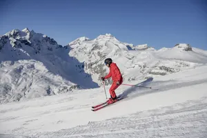 Skifahrer in roter Kleidung und schwarzem Helm fährt auf schneebedeckter Piste vor schneebedeckten Bergen in der Nähe des Hotel Mischun