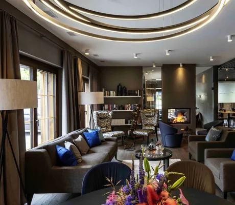 Lounge des Hotel Mischun mit Kamin, mehreren Sesseln und Sofas mit blauen Kissen, runden Deckenleuchten und einem Tisch mit Blumenarrangement im Vordergrund