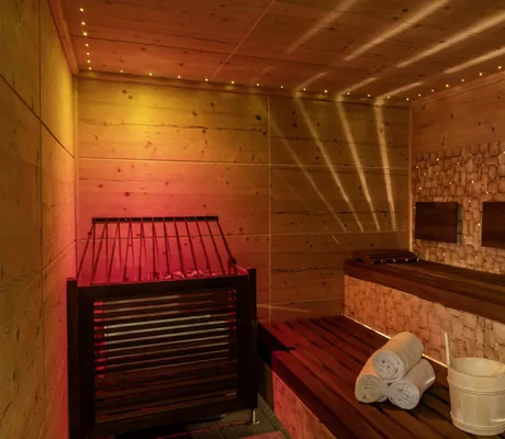 Sauna im Hotel Mischun mit Holzwänden, Holzbänken, Saunaofen, aufgerollten Handtüchern und Holzeimer mit Kelle