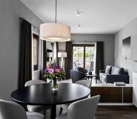 Moderner Wohnraum einer Suite des Hotel Mischun mit runder schwarzer Esstischplatte, grauen Stühlen, Küchenzeile, Sofa, Sesseln, großen Fenstern und Blumenstrauß auf dem Tisch