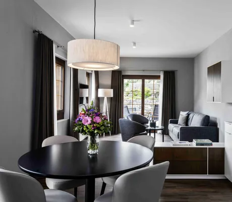 Moderner Wohnraum einer Suite des Hotel Mischun mit runder schwarzer Esstischplatte, grauen Stühlen, Küchenzeile, Sofa, Sesseln, großen Fenstern und Blumenstrauß auf dem Tisch