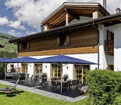 Terrasse und Fassade des Hotel Mischun, blaue Sonnenschirme, Gartenmöbel und Liegestuhl auf grünem Rasen vor bewaldeten Hügeln unter blauem Himmel mit Wolken