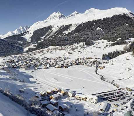 Verschneite Dorflandschaft in einem Tal in der Nähe des Hotel Mischun, umgeben von bewaldeten und schneebedeckten Bergen unter blauem Himmel