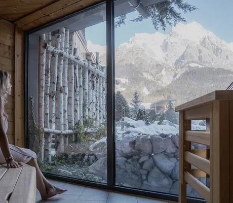 blonde Frau sitzt in Handtuch gewickelt in der Sauna der Riederalm und schaut auf die verschneiten Berge