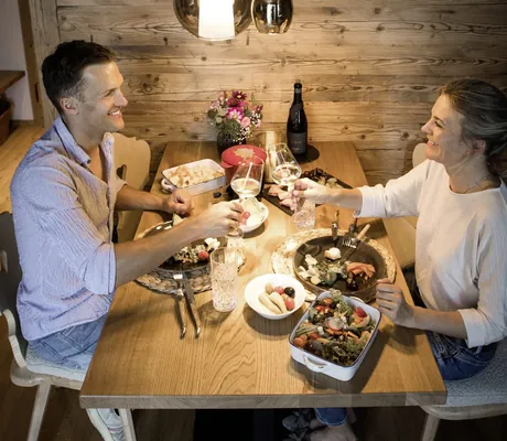 Zwei Personen sitzen an einem gedeckten Holztisch der Alpzitt-Chalets beim Abendessen und stoßen mit Weingläsern an, darüber hängen drei unterschiedlich große Pendelleuchten