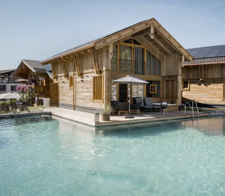 Neues Chalet der Alpzitt-Chalets mit Terrasse und Sonnenschirm direkt am klaren Naturbadesee unter blauem Himmel