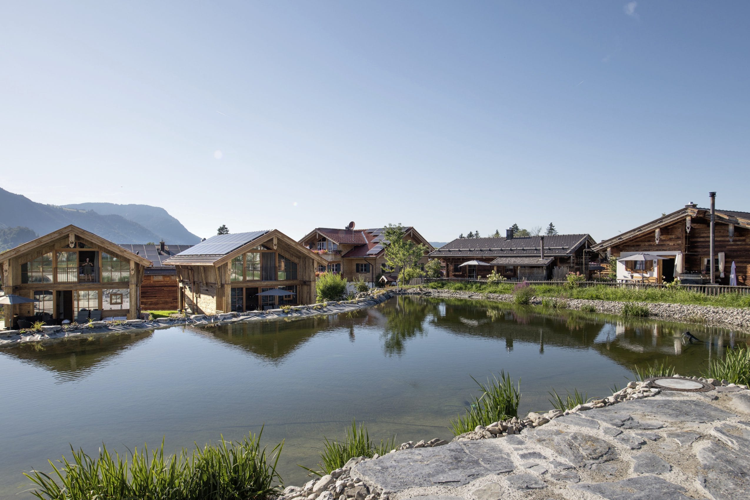 Alpzitt-Chalets am idyllischen Mühlenweiher mit Bergen im Hintergrund unter klarem Himmel