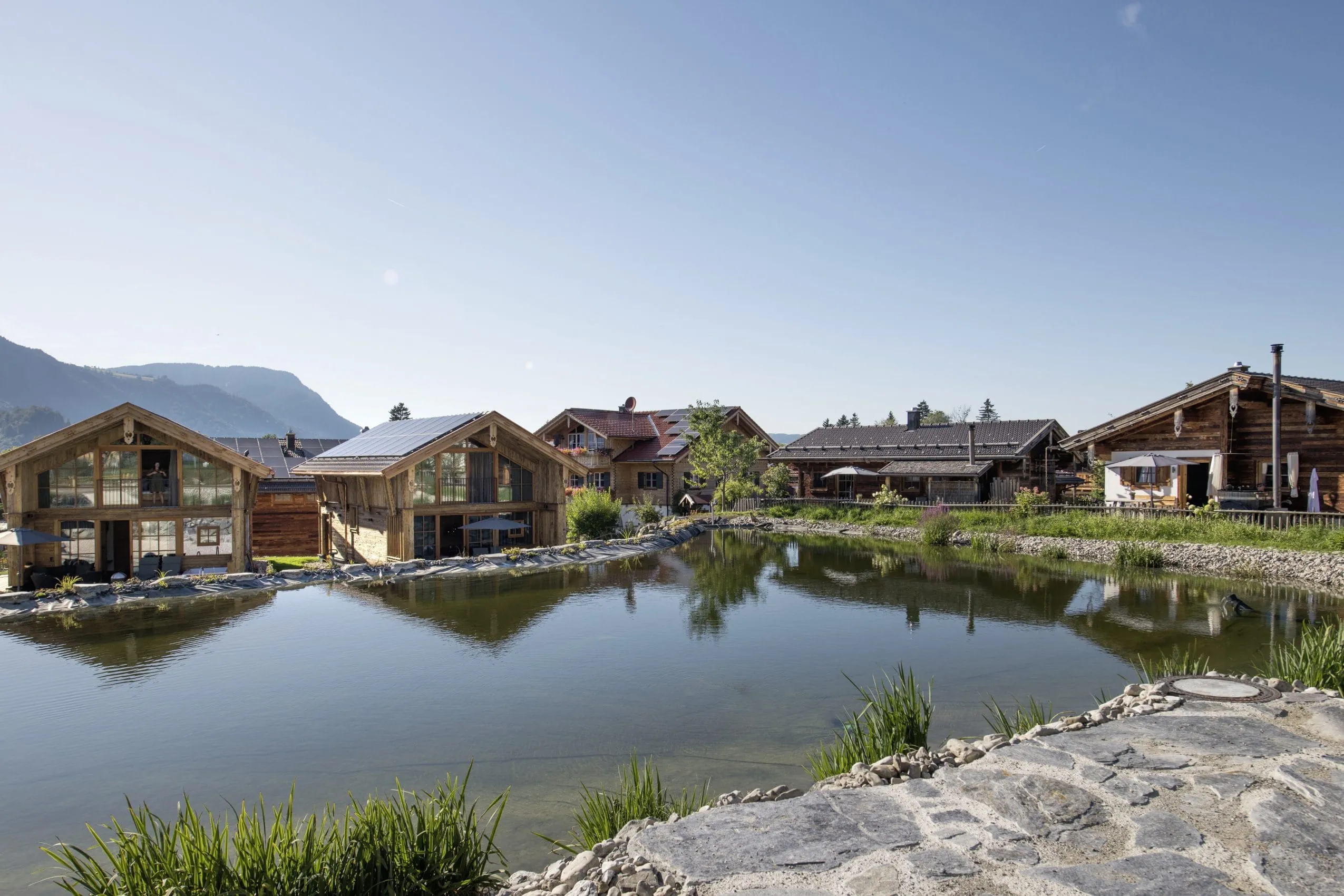 Alpzitt-Chalets am idyllischen Mühlenweiher mit Bergen im Hintergrund unter klarem Himmel