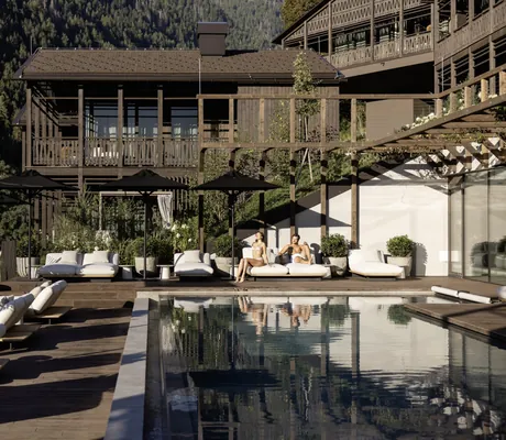 Paar liegt in Badekleidung auf den weißen Polstern am Beckenrand des Outdoorpools in den Amonti Chalets