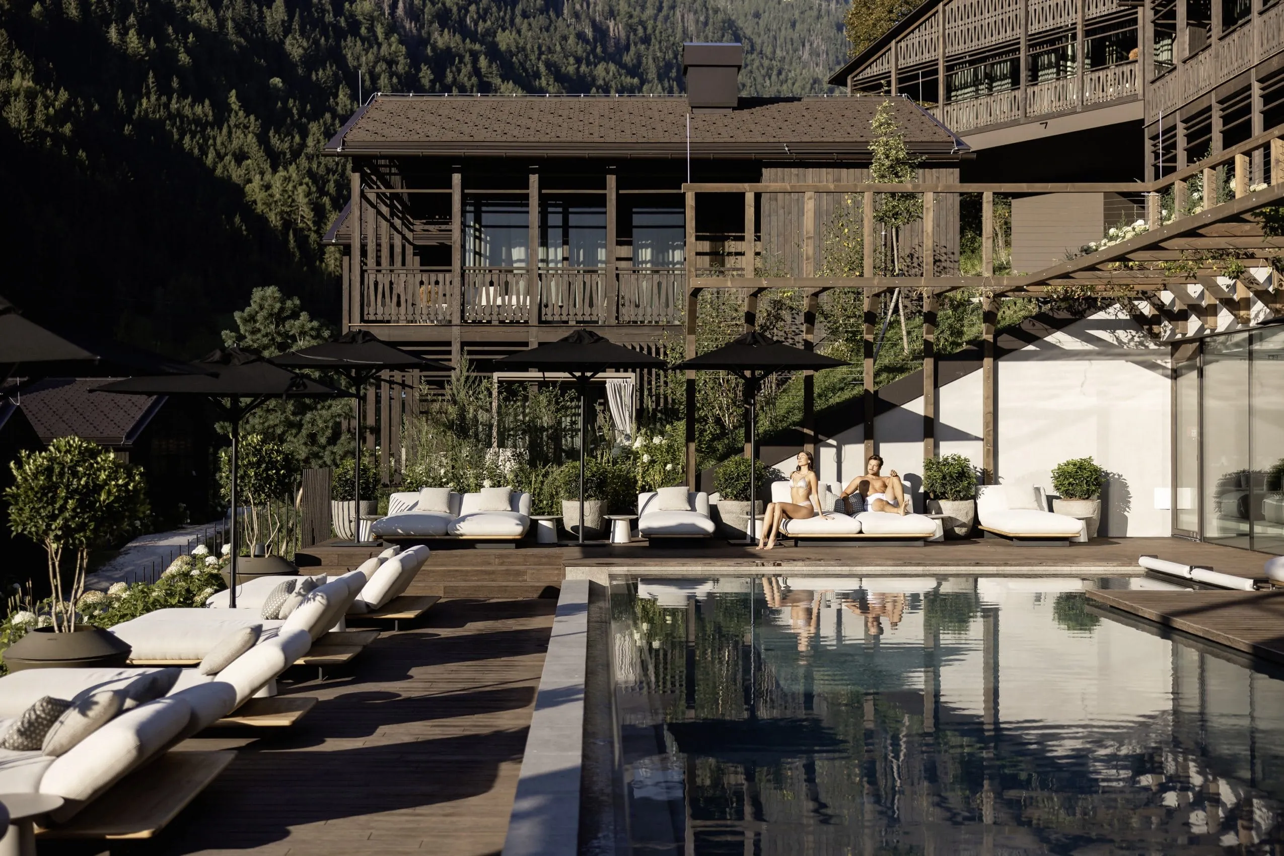 Paar liegt in Badekleidung auf den weißen Polstern am Beckenrand des Outdoorpools in den Amonti Chalets