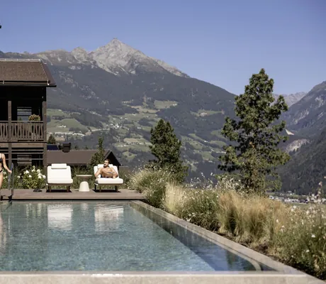 Frau in Bikini geht in den Outdoorpool der Amonti Chalets und Mann liegt auf der weißen Sonnenliege