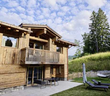 Außenansicht eines der Ziegelberg Chalets mit Balkon und Terrasse, davor zwei Liegestühle mit Sonnenschirm auf Rasen, im Hintergrund Wiese und hohe Nadelbäume