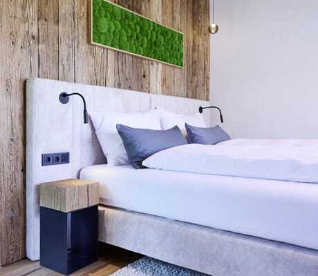 Modernes Schlafzimmer mit hellgrauem Polsterbett, weißer Bettwäsche, dunkelgrauen Kissen, Holzverkleidung an der Wand und grünem Wandbild in den Ziegelberg Chalets
