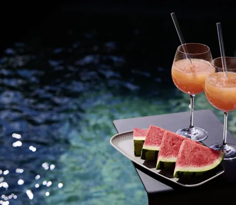 Zwei Gläser mit orangem Getränk und Glasstrohhalm neben vier Wassermelonenstücken auf einem schwarzen Tisch am Poolrand der Ziegelberg Chalets