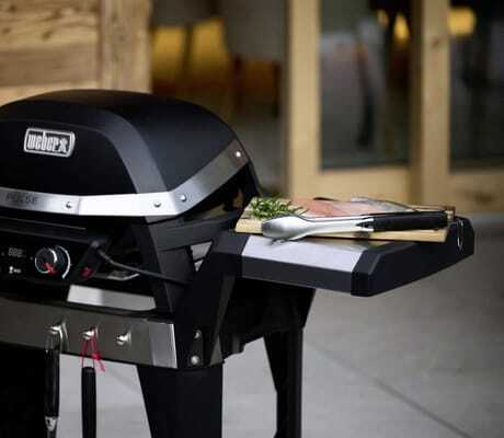 Schwarzer Weber-Gasgrill der Ziegelberg Chalets mit geschlossenem Deckel, zwei Drehreglern und seitlicher Ablage mit Grillzange, Fisch und Kräutern