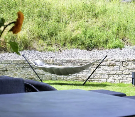 Beige Hängematte auf schwarzem Metallgestell vor einer Steinmauer und grünem Gras in den Ziegelberg Chalets, im Vordergrund unscharfe Blumen in Vase