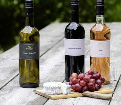 Drei Weinflaschen Weißwein, Rotwein und Rosé der Ziegelberg Chalets auf Holztisch, davor Käse und rote Trauben