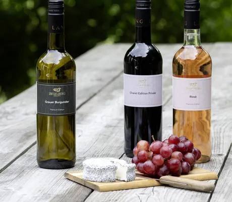 Drei Weinflaschen Weißwein, Rotwein und Rosé der Ziegelberg Chalets auf Holztisch, davor Käse und rote Trauben