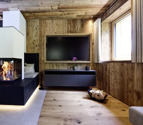 Moderner Kamin mit Glasfront, schwarzer Sockel, Holzwand und großer Fernseher in hellem Wohnzimmer mit Holzboden in den Ziegelberg Chalets