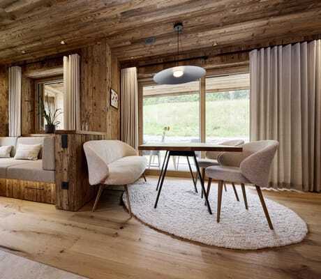 Wohn- und Essbereich in den Ziegelberg Chalets mit Holzwänden und -boden, Ecksofa mit Kissen, Tisch, Stühle und Sitzbank auf rundem Teppich, große Fenster mit Vorhängen