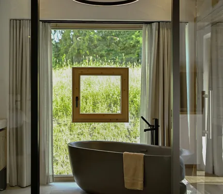 Freistehende, dunkelgraue Badewanne mit Handtuch vor quadratischem Fenster mit Vorhängen und runder Deckenlampe in den Ziegelberg Chalets