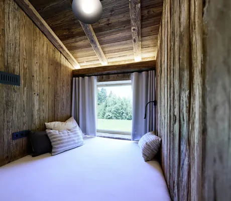 Kleiner Schlafbereich in den Ziegelberg Chalets mit Holzwänden, weißer Matratze, Kissen und Fenster mit Blick auf grüne Wiese und Bäume