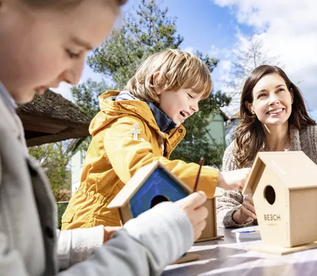 dunkelhaarige Frau bastelt mit Kind in gelber Jacke im BEECH Resort Fleesensee ein Vogelhäuschen