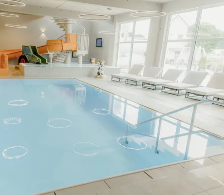 Indoorpool, orangefarbene Wasserrutsche, weiße Liegen und große Fenster im Familienhotel Friedrichshof